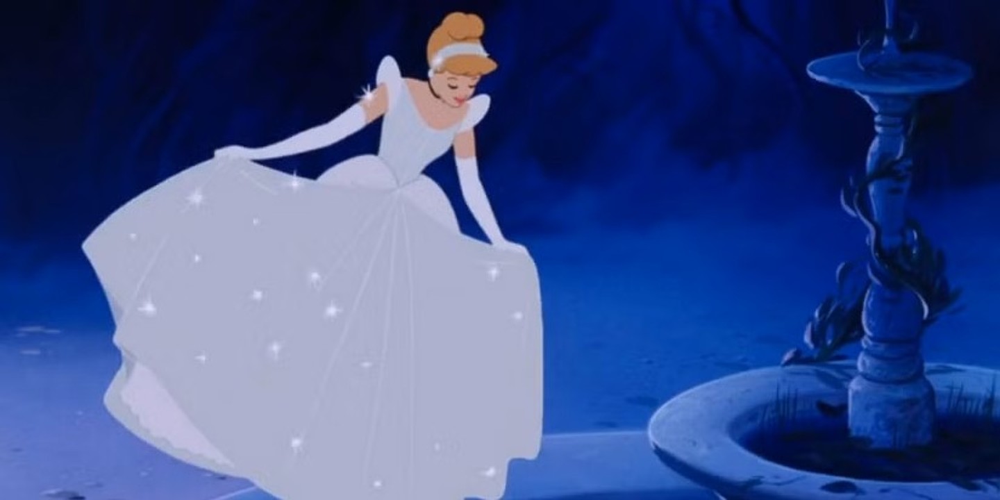 L'image a un fond bleu et noir. Il y a une fontaine à droite de l'image. La princesse Cendrillon tient sa robe dans ses mains et s'observent dans l'eau de la fontaine. Sa robe est de couleur argente, presque bleue. Elle porte des gants de la même couleur ainsi qu'une passe sur sa chevelure attachée en chignon.
