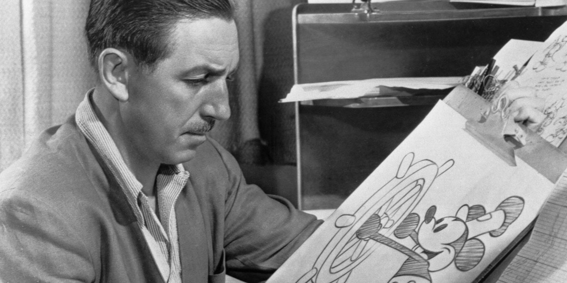 L'image est en noir et blanc. Monsieur Walt Disney est en train de dessiner Mickey Mouse avec la roue de son bateau « Steamboat »